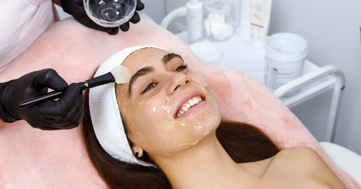 Smiling woman receiving Chemical Peels in Henderson, NV, as an aesthetician applies a gel mask in a spa.