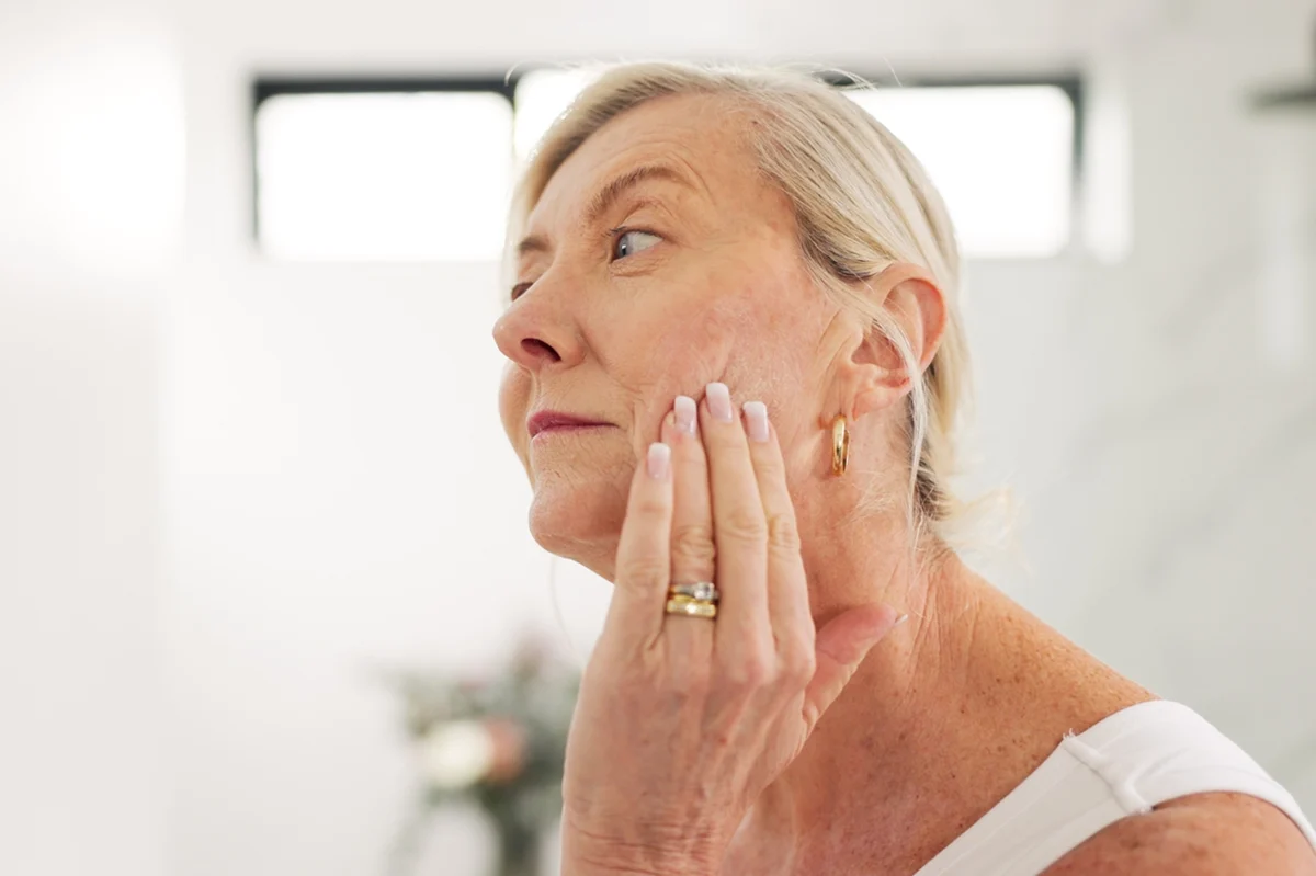 Older woman touching her cheek, showing visible acne scars and skin texture.